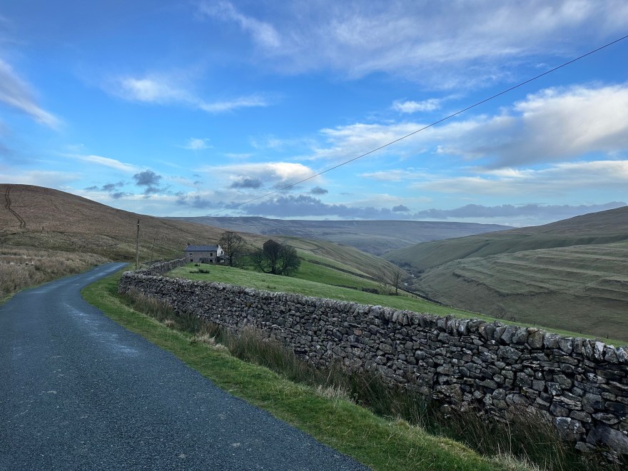 A road leading toward rolling hills with a small house visible in the distance.